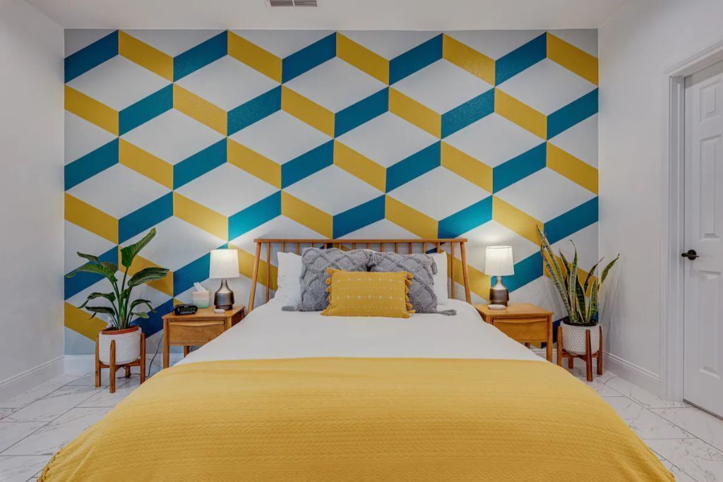 Una habitación de hotel con una pared de diseño geométrico, una cama con cubrecama amarillo, dos mesitas de noche con lámparas y dos plantas en macetas.