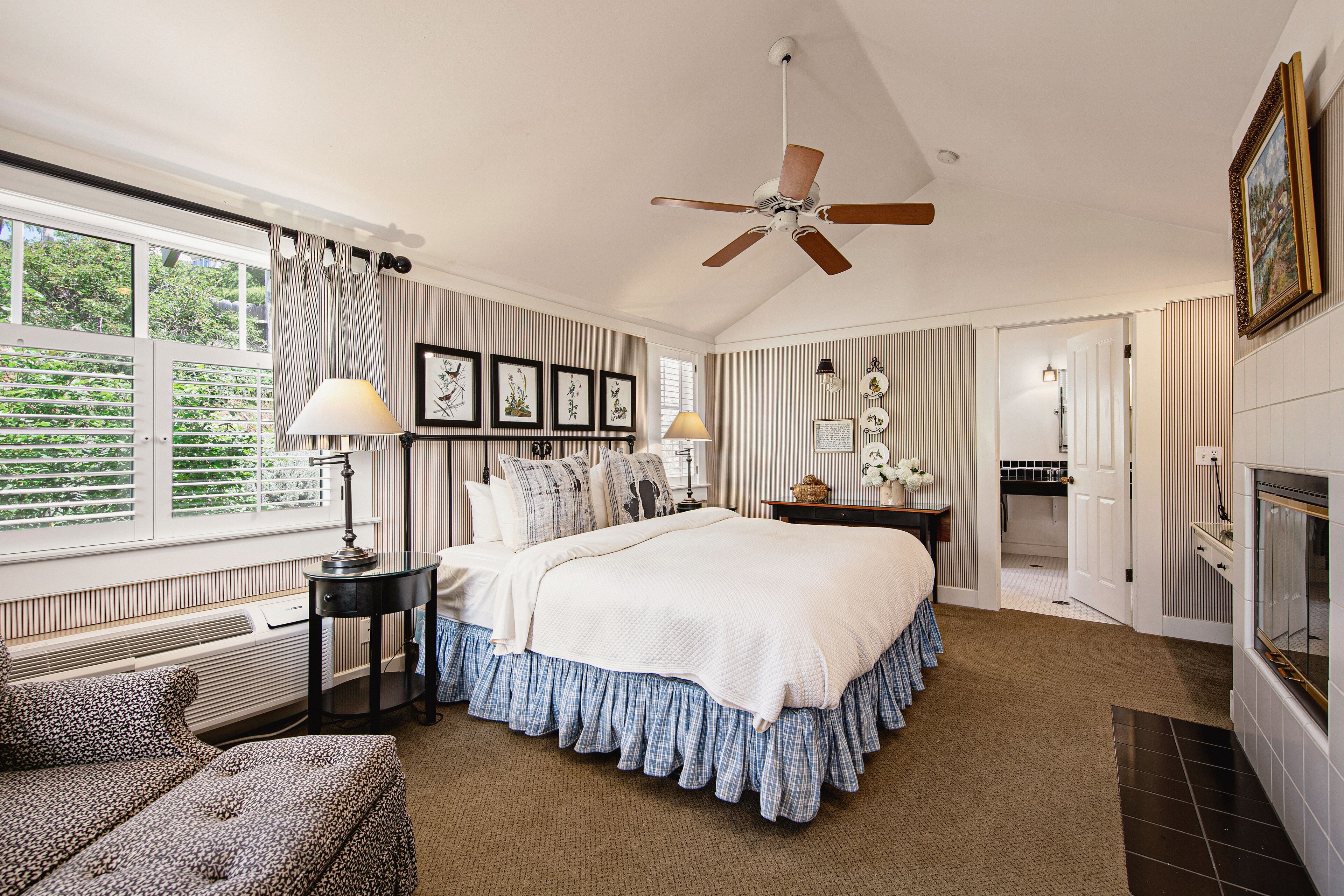 A bedroom with a large bed, a ceiling fan, a fireplace, and a view of greenery outside.