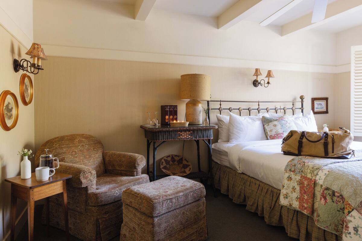 A cozy bedroom with a bed, a chair, a table with a lamp, and a small side table.