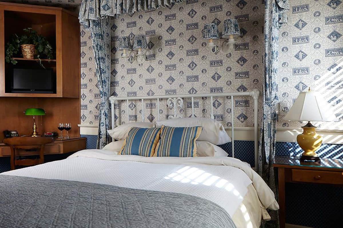 A hotel room with a bed, a wooden cabinet with a television, a desk with a lamp, and patterned wallpaper.