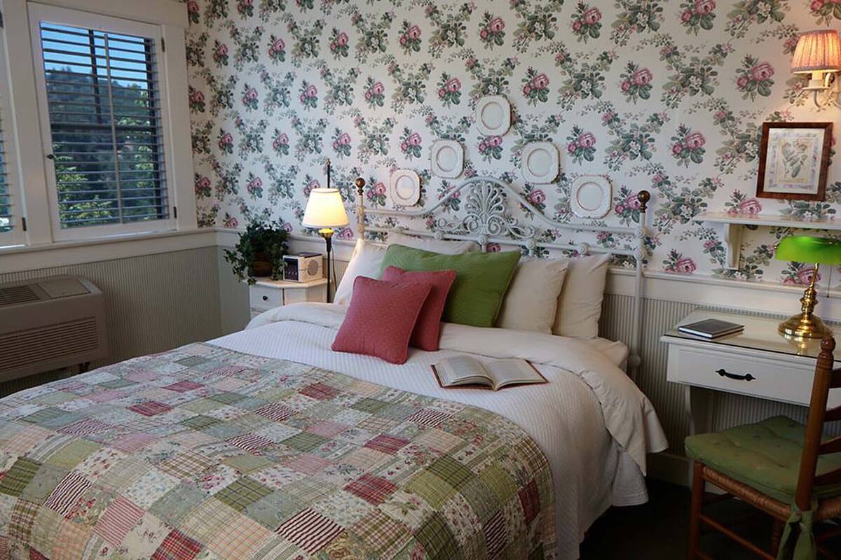 A bedroom with a floral wallpaper, a bed with a quilt, bedside lamps, a chair, and a desk with a lamp.