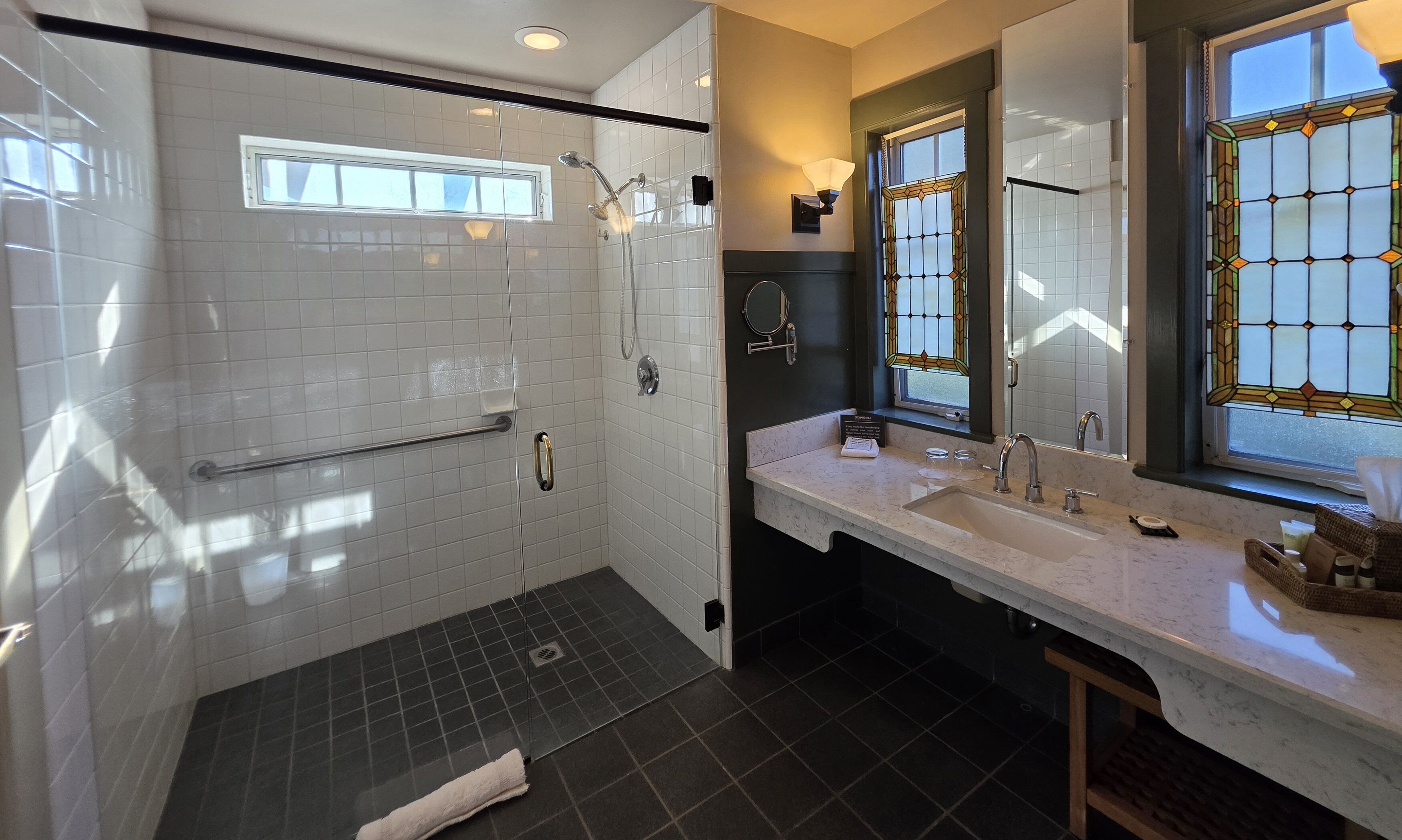 A modern bathroom with a walk-in shower, a vanity with a large mirror, and a window with stained glass.