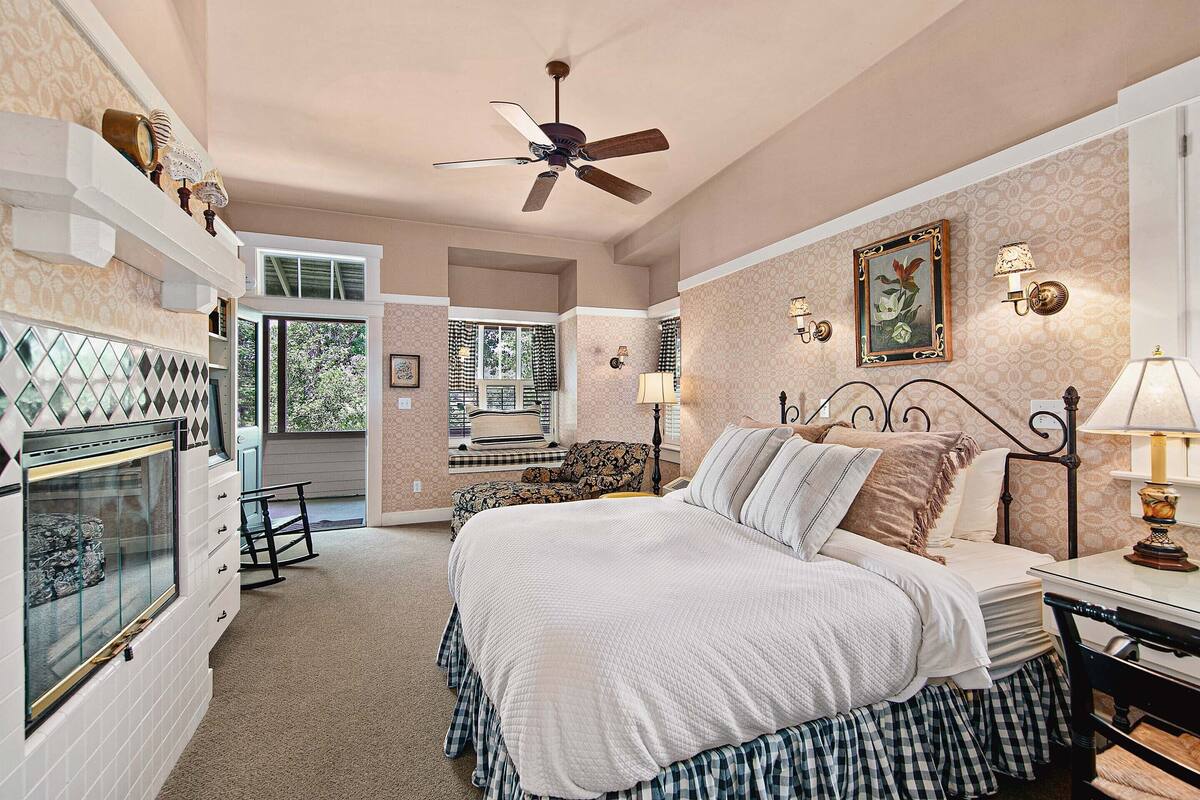 A bedroom with a bed, a ceiling fan, a fireplace, and a window with a view of the outdoors.
