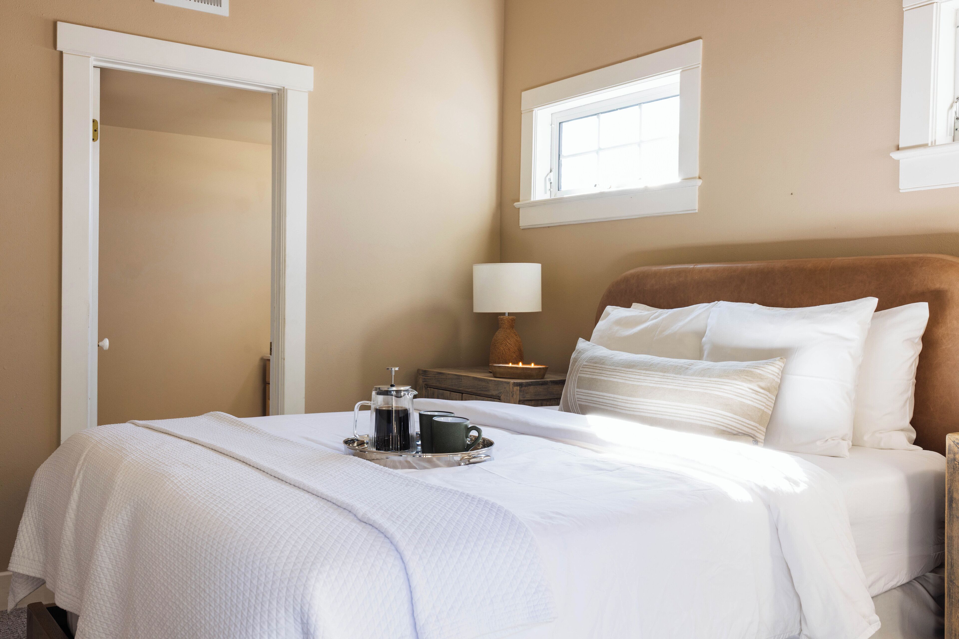 A neatly made bed with white linens, a bedside table with a lamp, and a tray with a coffee pot and cups.