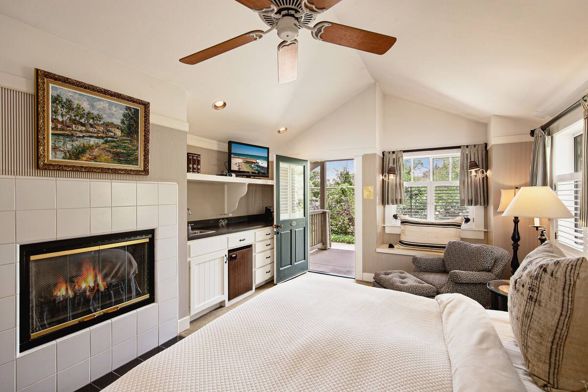 A hotel room with a fireplace, a ceiling fan, a television, a sofa, and a view of the outdoors.