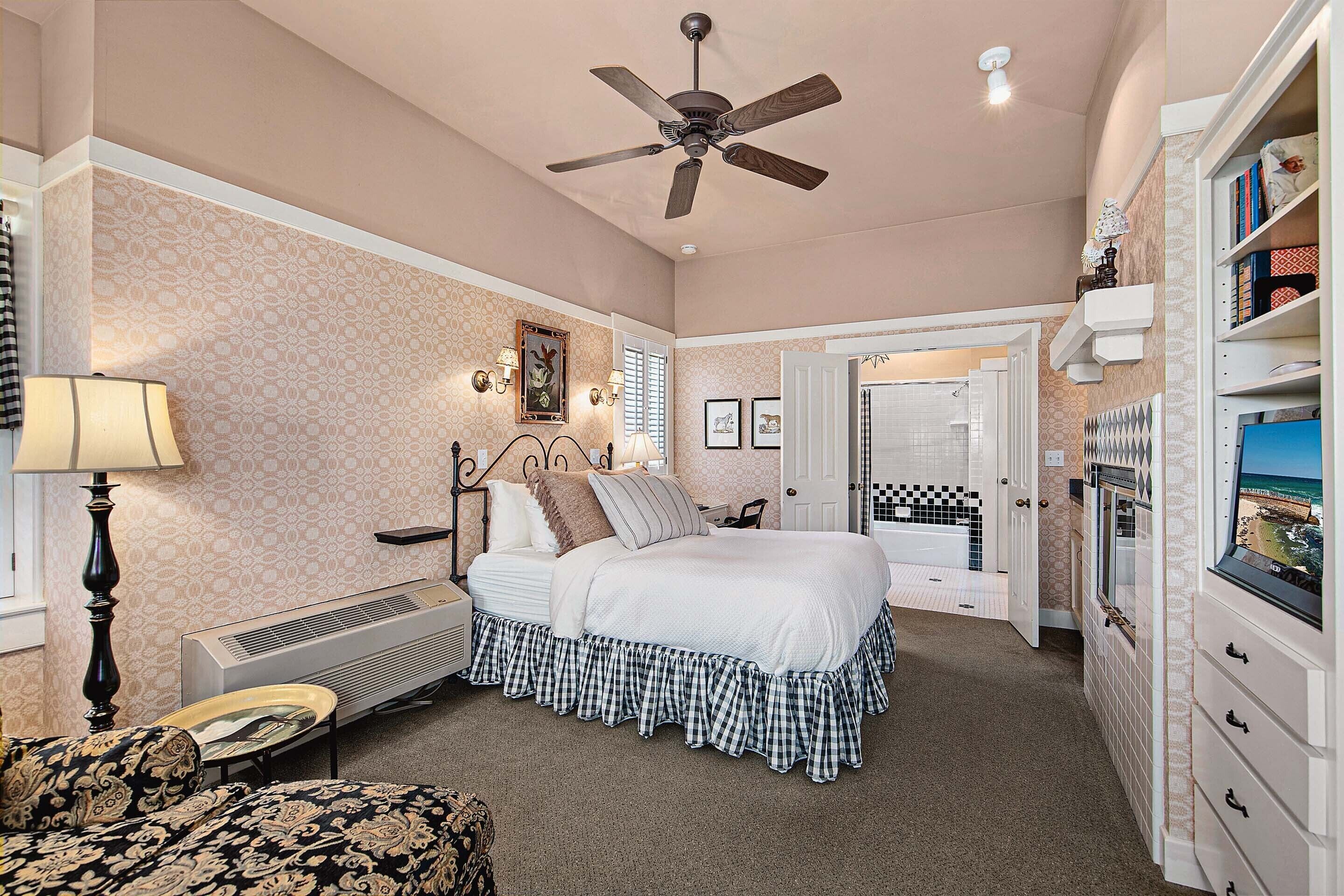A bedroom with a bed, a ceiling fan, a radiator, a lamp, and a view of the outside through a window.