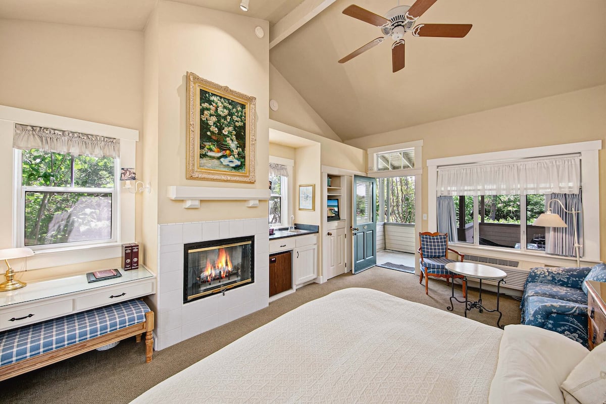 A spacious bedroom with a fireplace, a large bed, a ceiling fan, and a view of the outdoors.