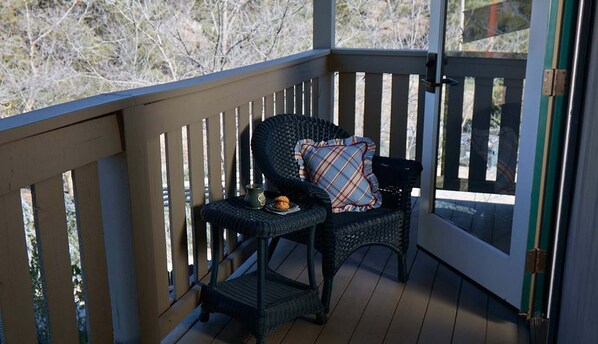 A covered outdoor seating area with a wooden deck, a wicker chair with a plaid cushion, and a small table with a mug and a pastry.