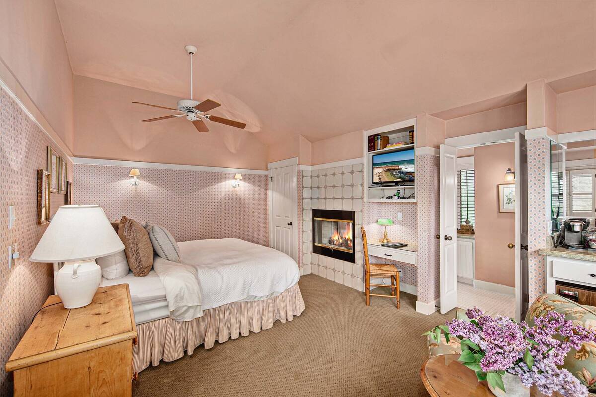 A bedroom with a bed, a desk, a chair, a fireplace, and a ceiling fan.