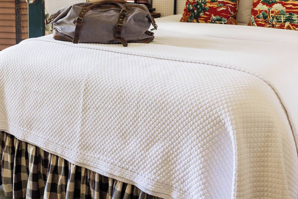 A neatly made bed with a quilted white cover, a brown bag on the bed, and a wall-mounted lamp.