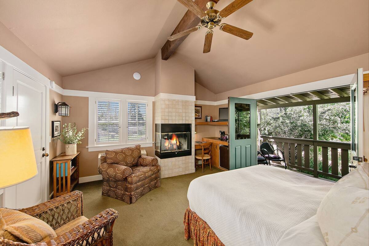A bedroom with a large bed, a fireplace, a balcony, wicker furniture, and a ceiling fan.