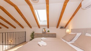A bedroom with a sloped ceiling, wooden beams, a bed with beige bedding, a bedside table with a mirror, a hanging plant, and a view of the outside through a skylight.