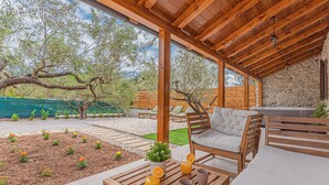 A covered outdoor seating area with wooden furniture, a stone wall, and a garden with flowers and a tree.