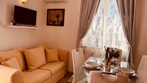 Una sala de estar con un sofá color beige, un televisor de pantalla plana, un equipo de aire acondicionado, un cuadro y una zona de comedor con una mesa dispuesta para dos.
