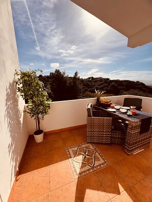 A balcony with a dining table, chairs, a potted plant, and a view of the surrounding landscape.