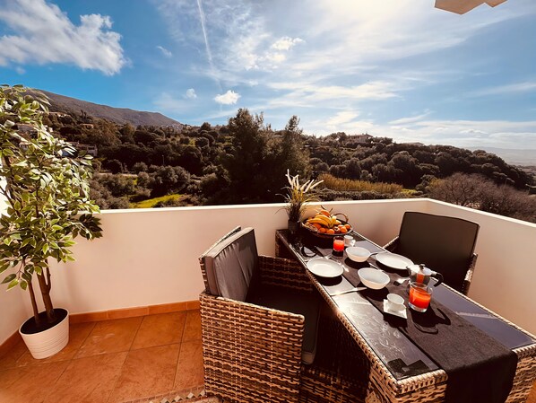 A balcony with a dining table set for two, surrounded by wicker chairs, a potted plant, and a view of a mountainous landscape.
