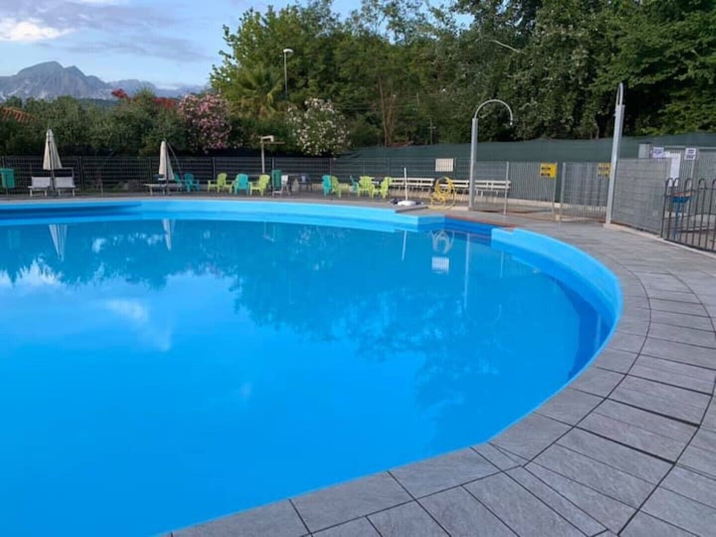 Ein Schwimmbecken mit einer gefliesten Kante, umgeben von grünen Stühlen und Sonnenschirmen, mit Bäumen und Bergen im Hintergrund.