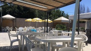 An outdoor patio area with white plastic tables and chairs, covered by a thatched roof.