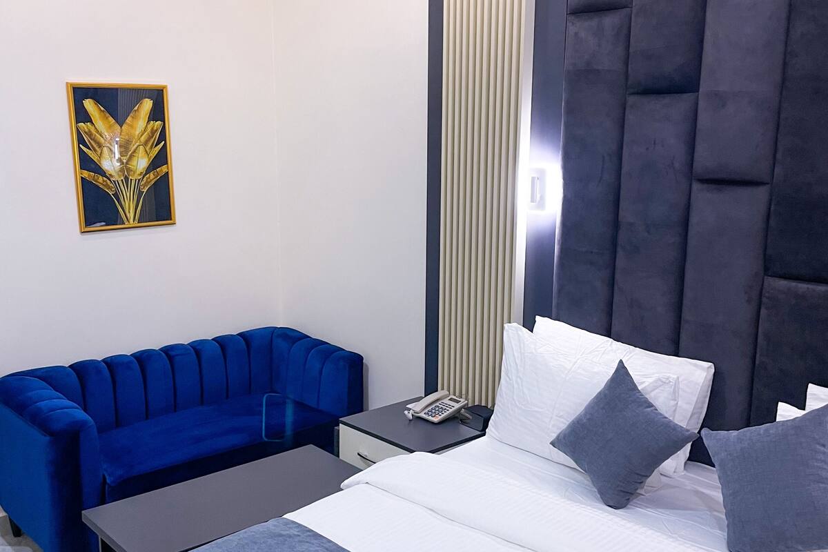 Una habitación de hotel moderna con una cama grande, un sofá azul, una mesita de noche con un teléfono y una lámpara de pared.
