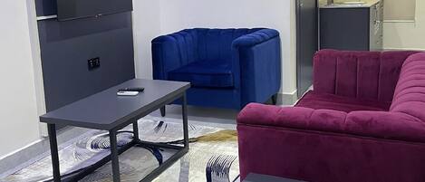 A modern living room with a flat-screen TV mounted on the wall, a blue sofa, a red sofa, and a small table with a remote control.