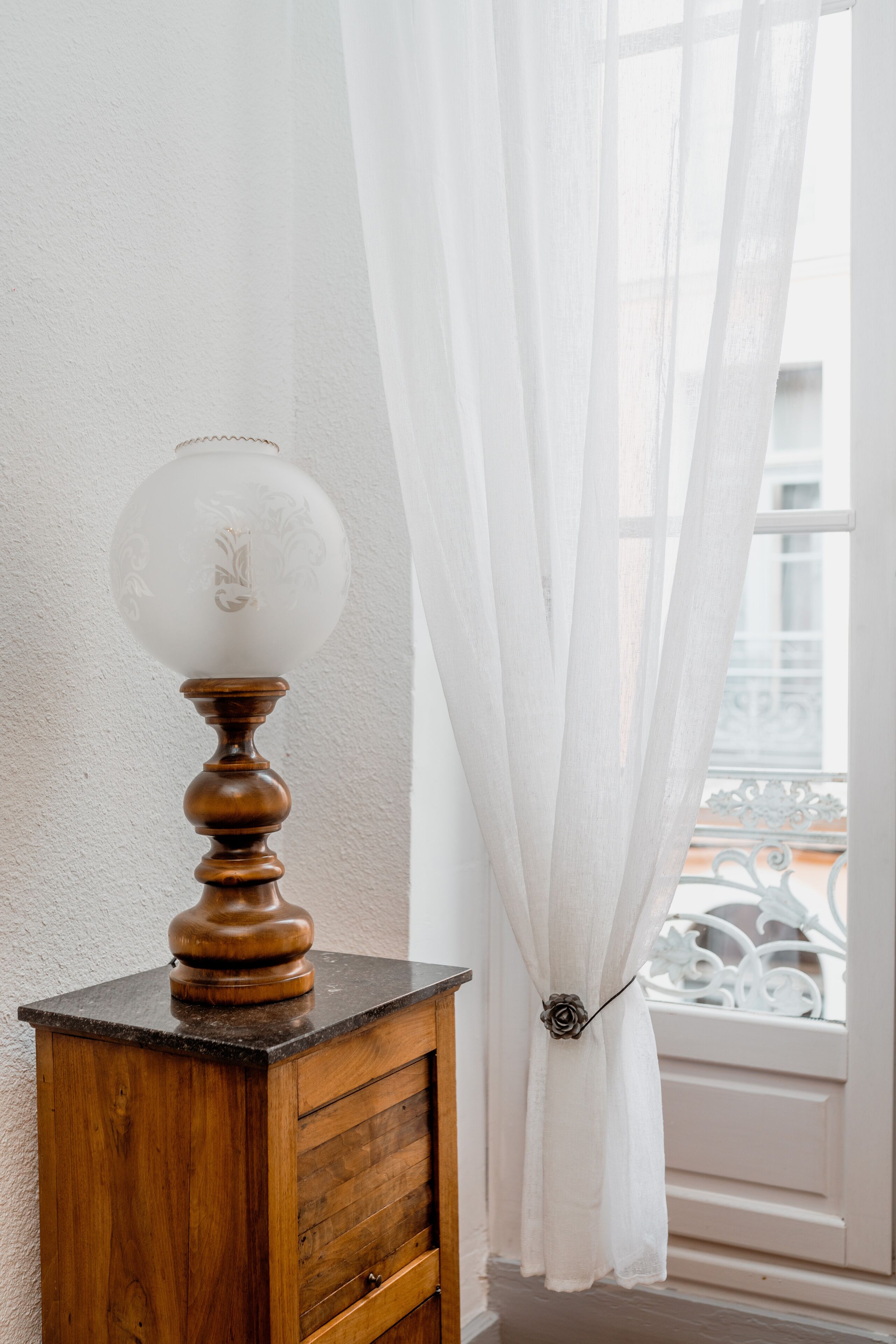 Un meuble en bois surmonté d’une lampe en verre dépoli, placé à côté d’une fenêtre ornée de rideaux blancs.