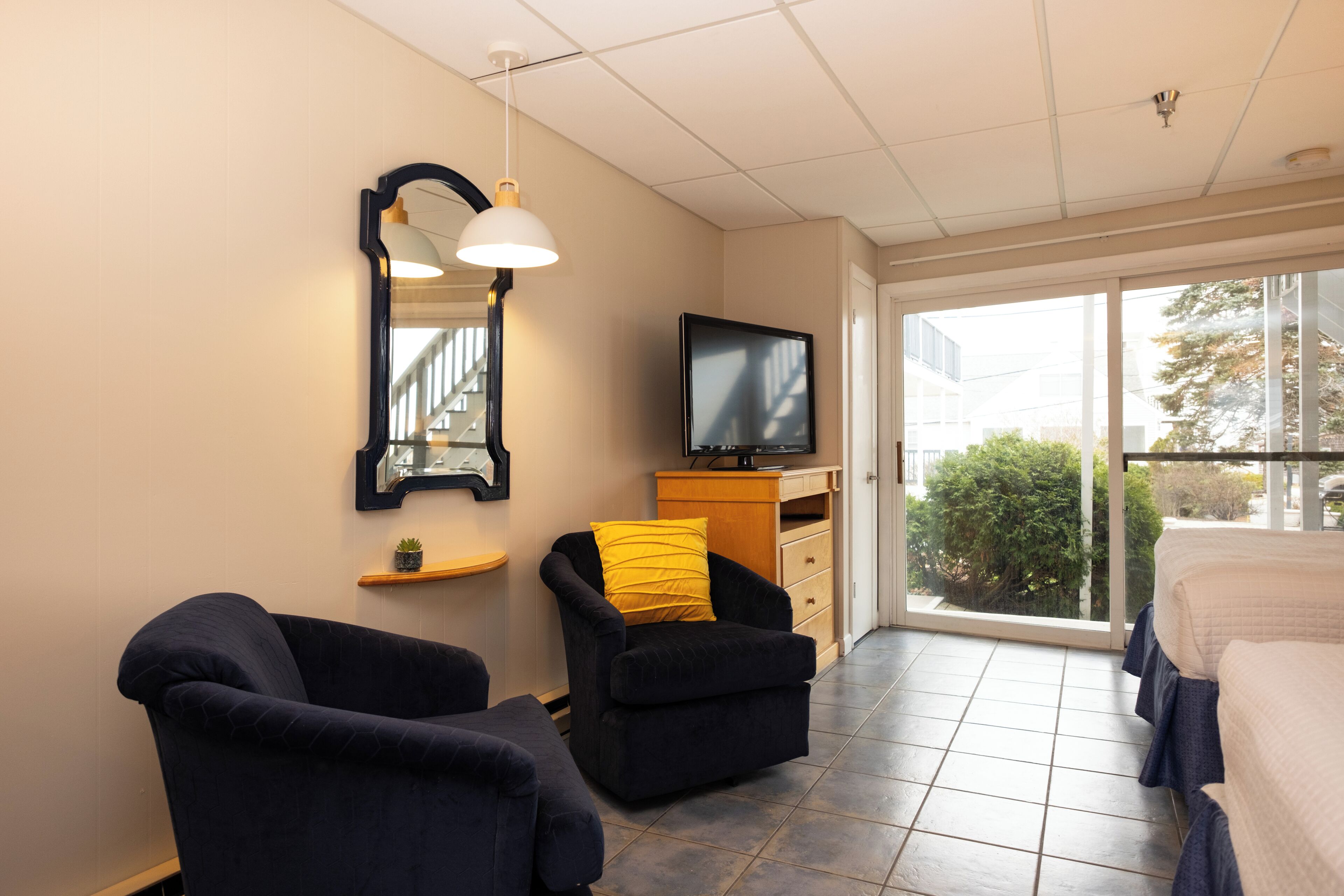 Habitación de hotel con un ventanal grande, un televisor de pantalla plana, dos sillones oscuros, una mesita con una planta y un cojín amarillo en uno de los sillones.