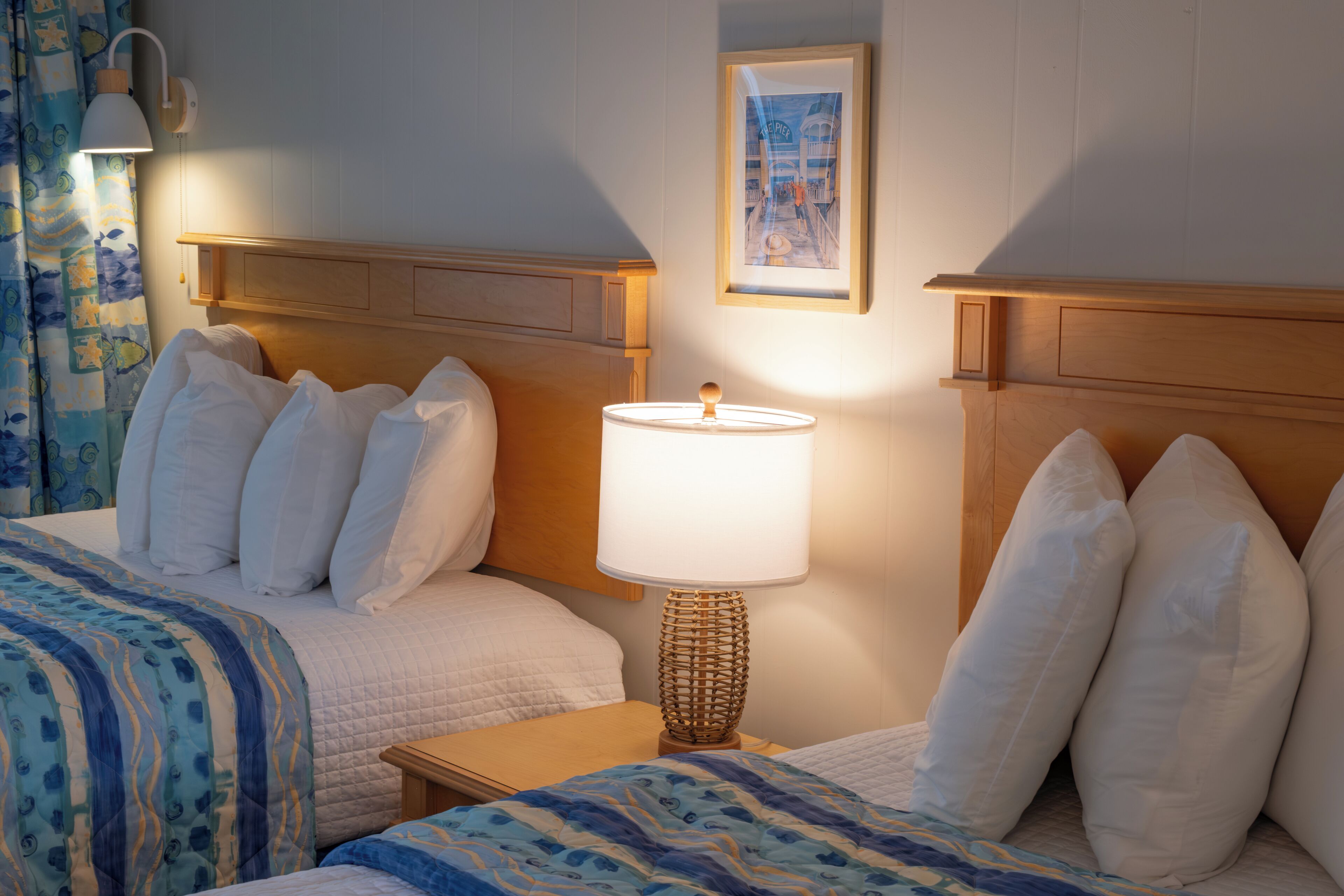 A hotel room with two beds, a bedside table with a lamp, a framed picture on the wall, and a curtain with a fish pattern.