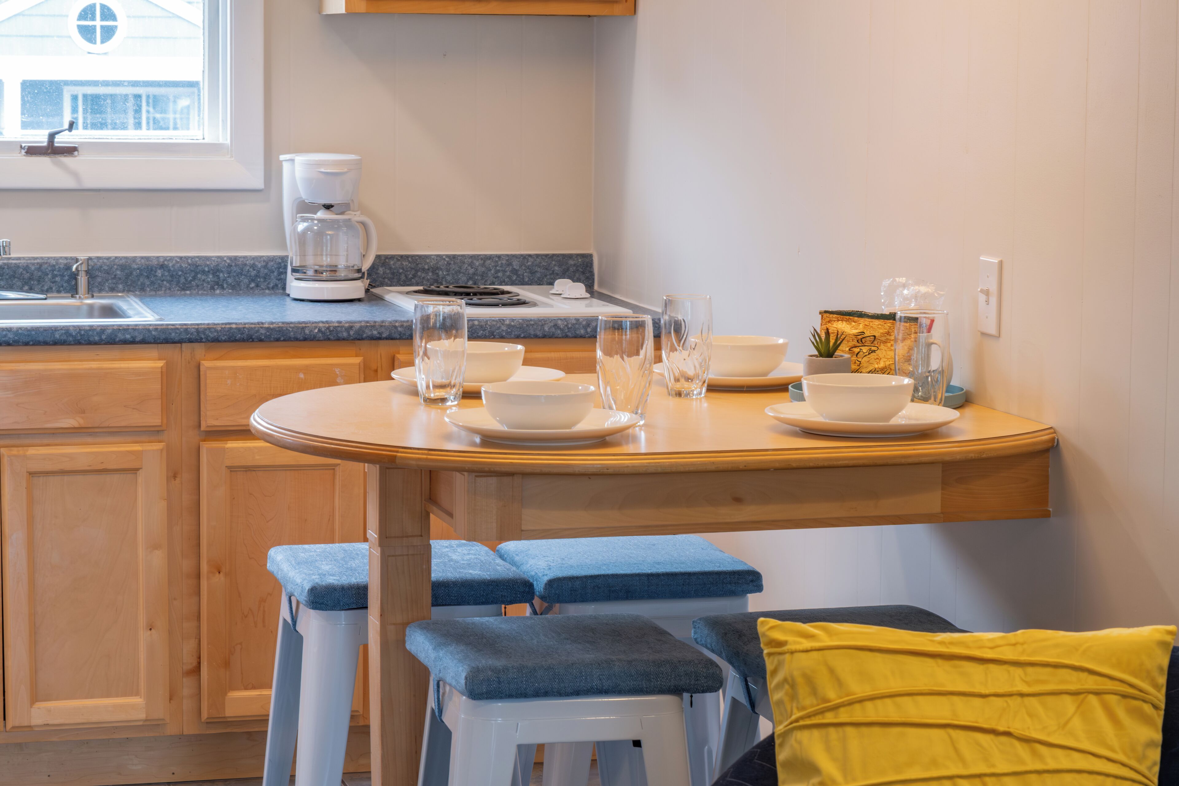 A kitchen with wooden cabinets, a countertop, a coffee maker, and a dining area with stools and a table set with bowls and glasses.