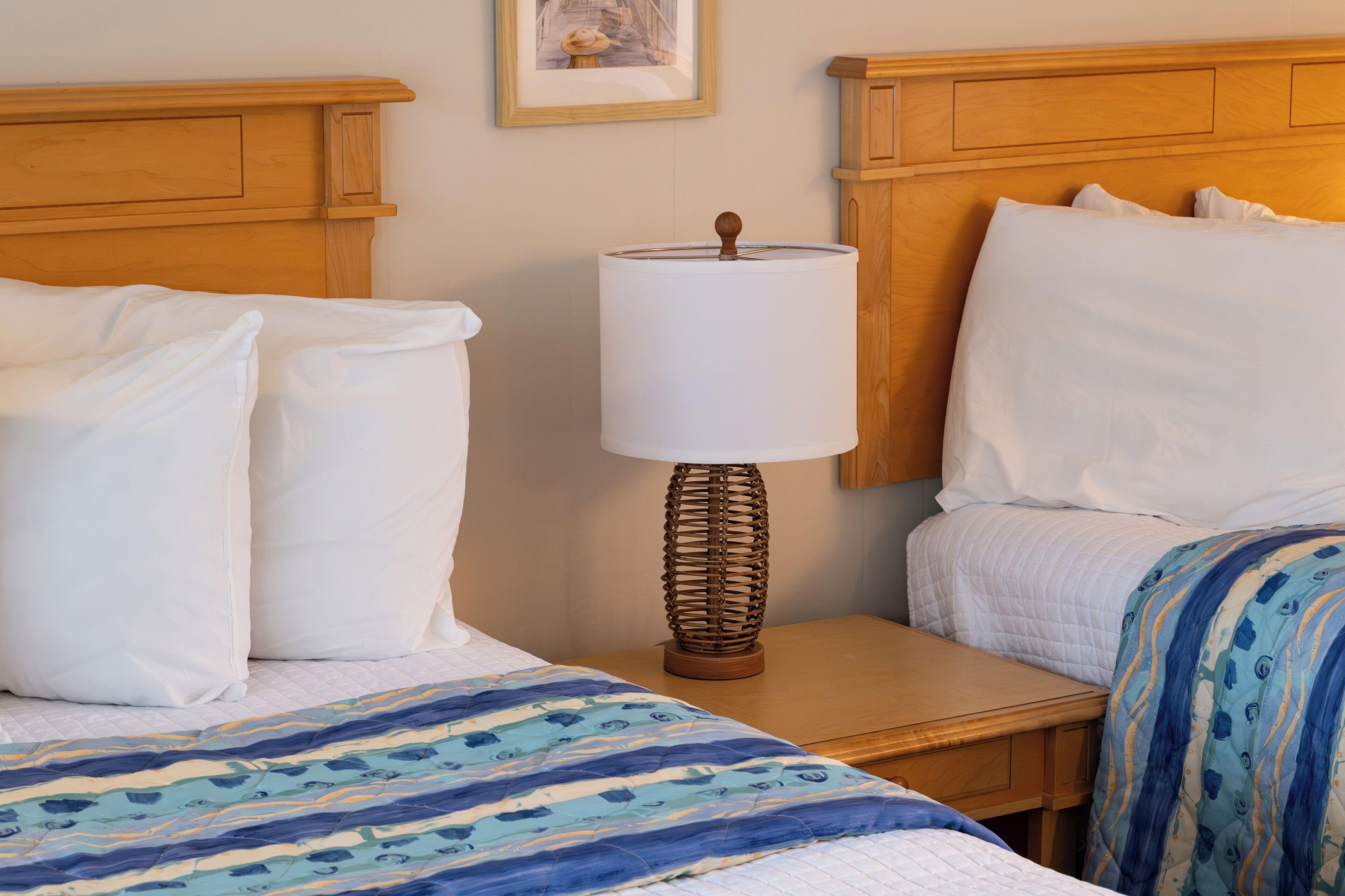 A hotel room with two beds, a bedside table with a lamp, and a framed picture on the wall.