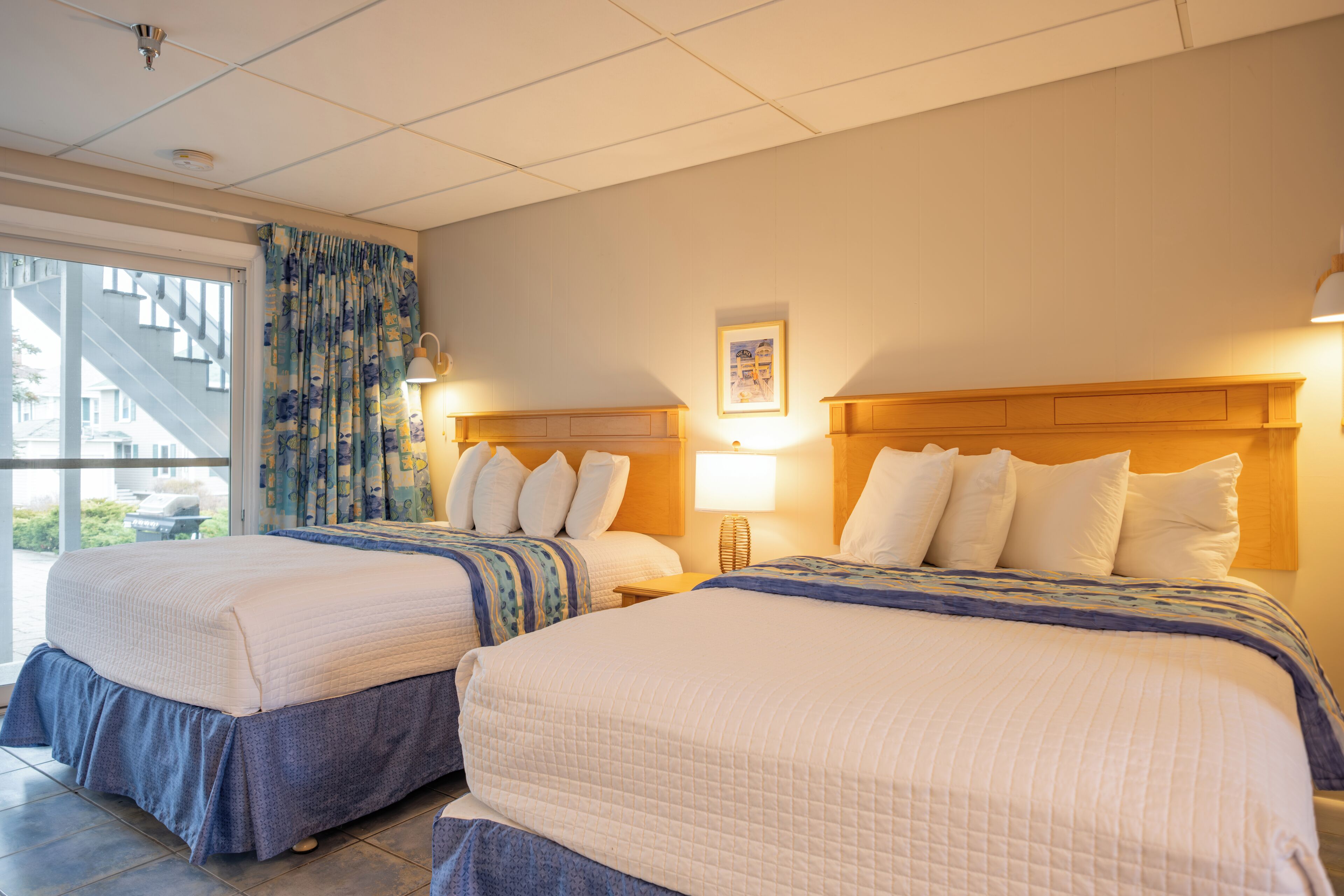 A hotel room with two beds, a large window with a view of a building, and a door leading outside.