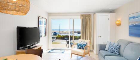 A modern living room with a television, a sofa, a dining table, and a view of the beach through the glass door.