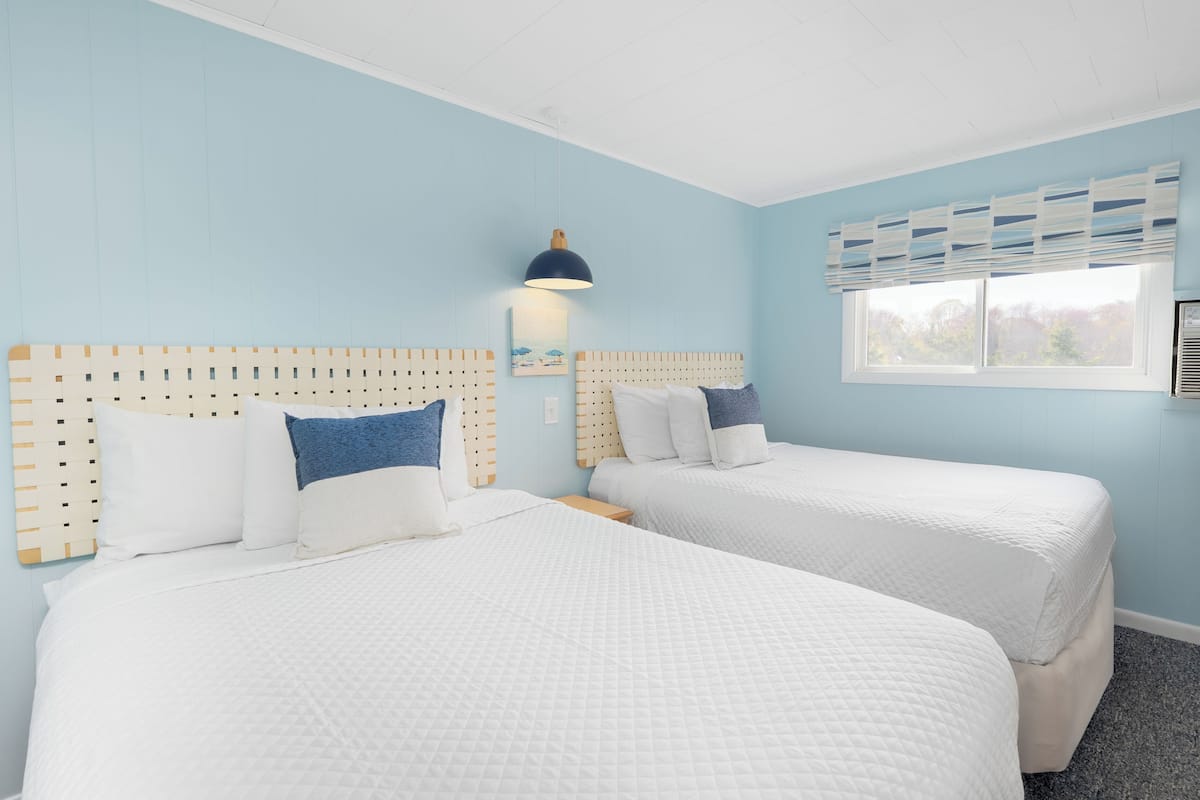 Een hotelkamer met twee bedden, een raam met zonwering, een wandairconditioner en een plafondlamp.