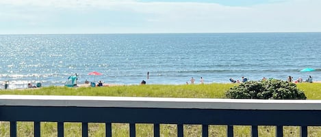 A beach with a clear view of the ocean, people relaxing, and a grassy area with a fence in the foreground.