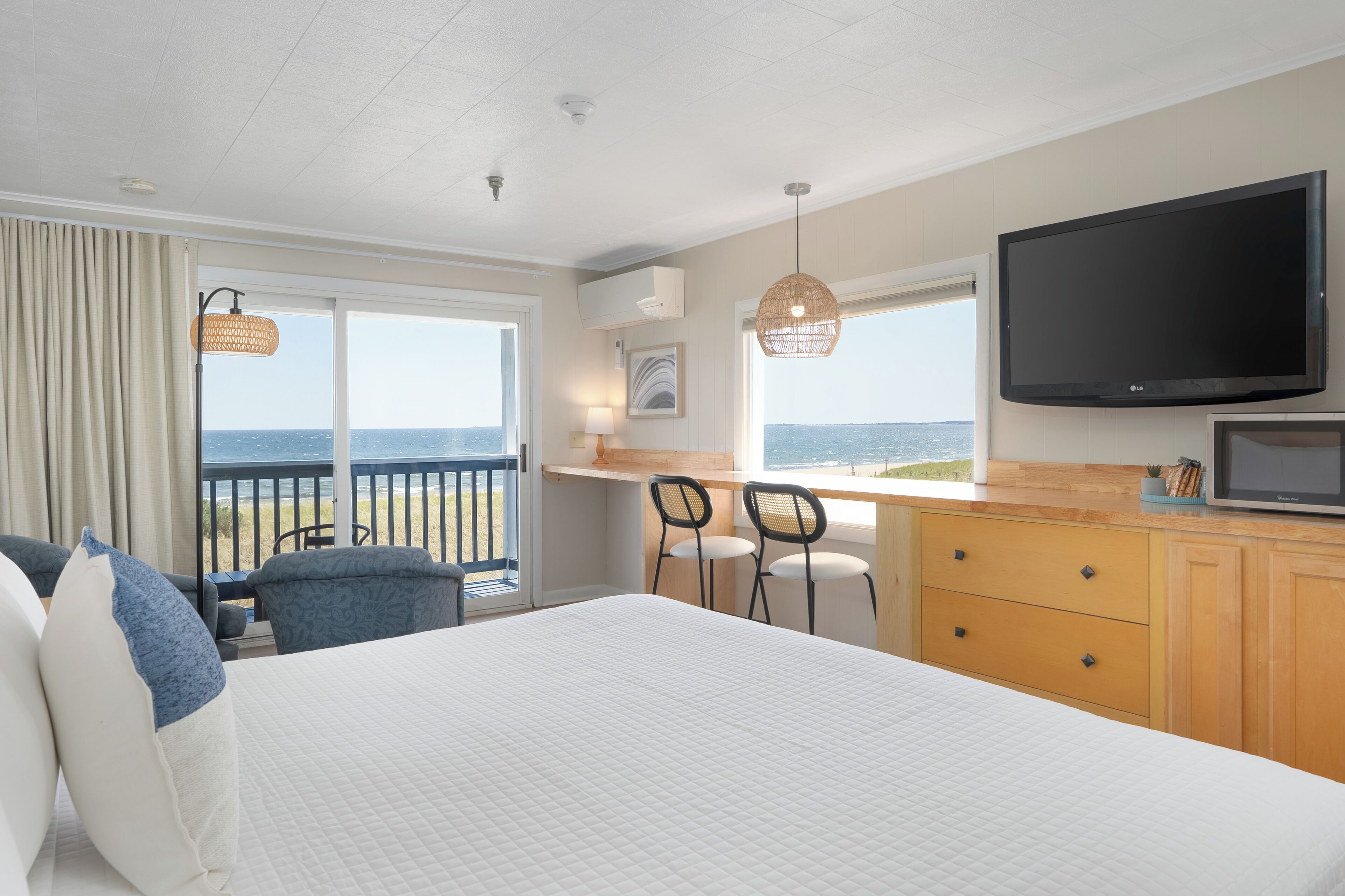 A hotel room with a bed, a desk, two chairs, a TV, and a view of the sea.