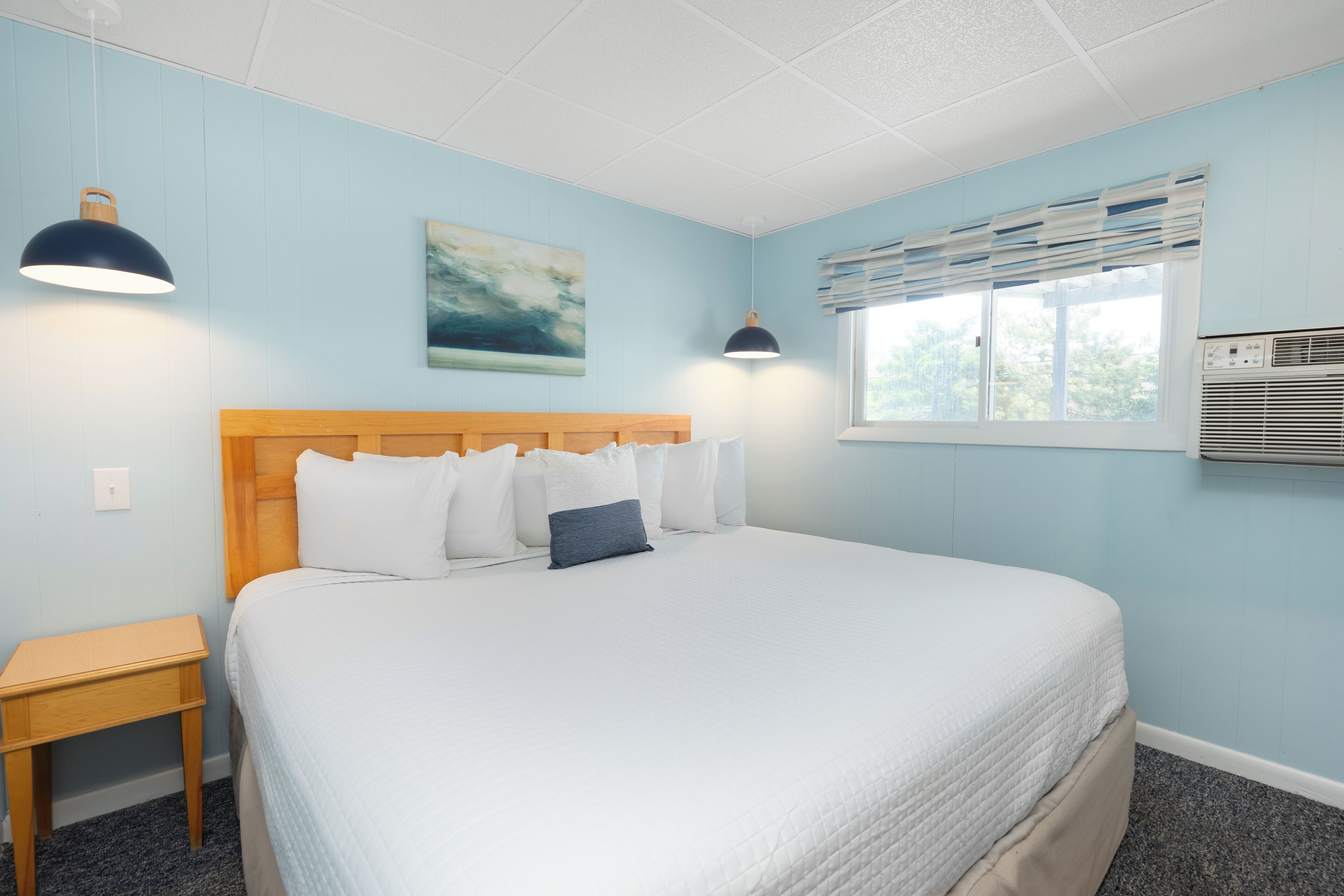 A neatly made bed with white bedding and a wooden headboard, a bedside table, a wall-mounted air conditioner, and a window with blinds.