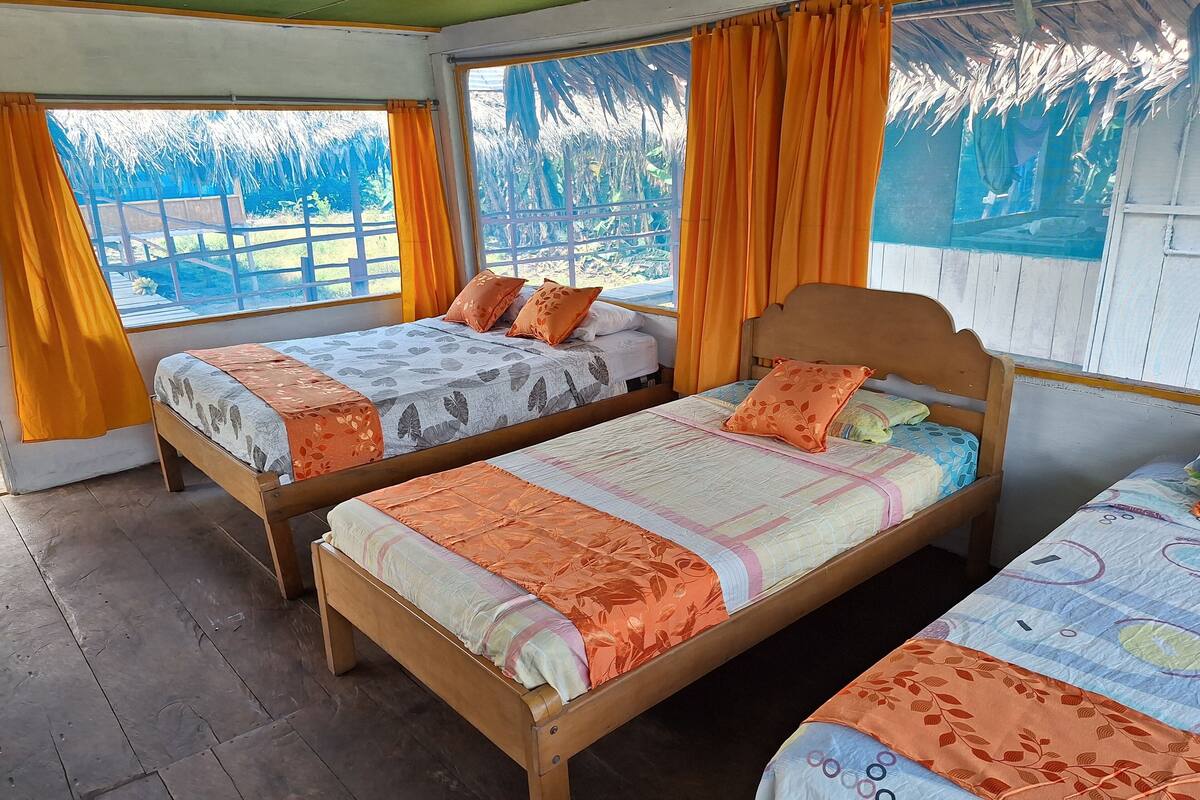 Two wooden beds with floral-patterned bedding, orange curtains, and a view of a thatched roof structure outside.