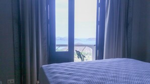 Una habitació amb un llit, una finestra amb cortines i vista al mar i a les muntanyes.