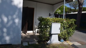 A covered outdoor area with a door, a small white utility box, and a hedge. - Angela Bungalow (Massa)