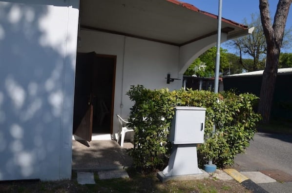 A covered outdoor area with a door, a small white utility box, and a hedge.
