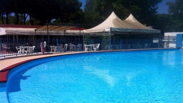 A swimming pool with a red edge, surrounded by a fence and a covered area with chairs. - Angela Bungalow (Massa)