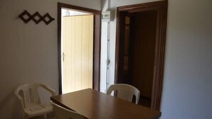 A room with a wooden table and two white plastic chairs, a wooden door, and a wall-mounted light switch. - Angela Bungalow (Massa)
