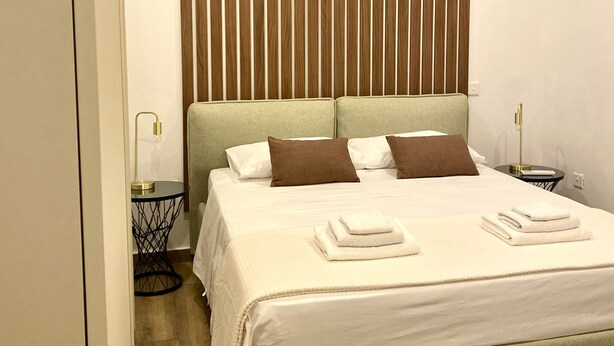A neatly arranged hotel room with a large bed, a wooden slat headboard, a bedside table with a lamp, and an air conditioning unit on the wall.