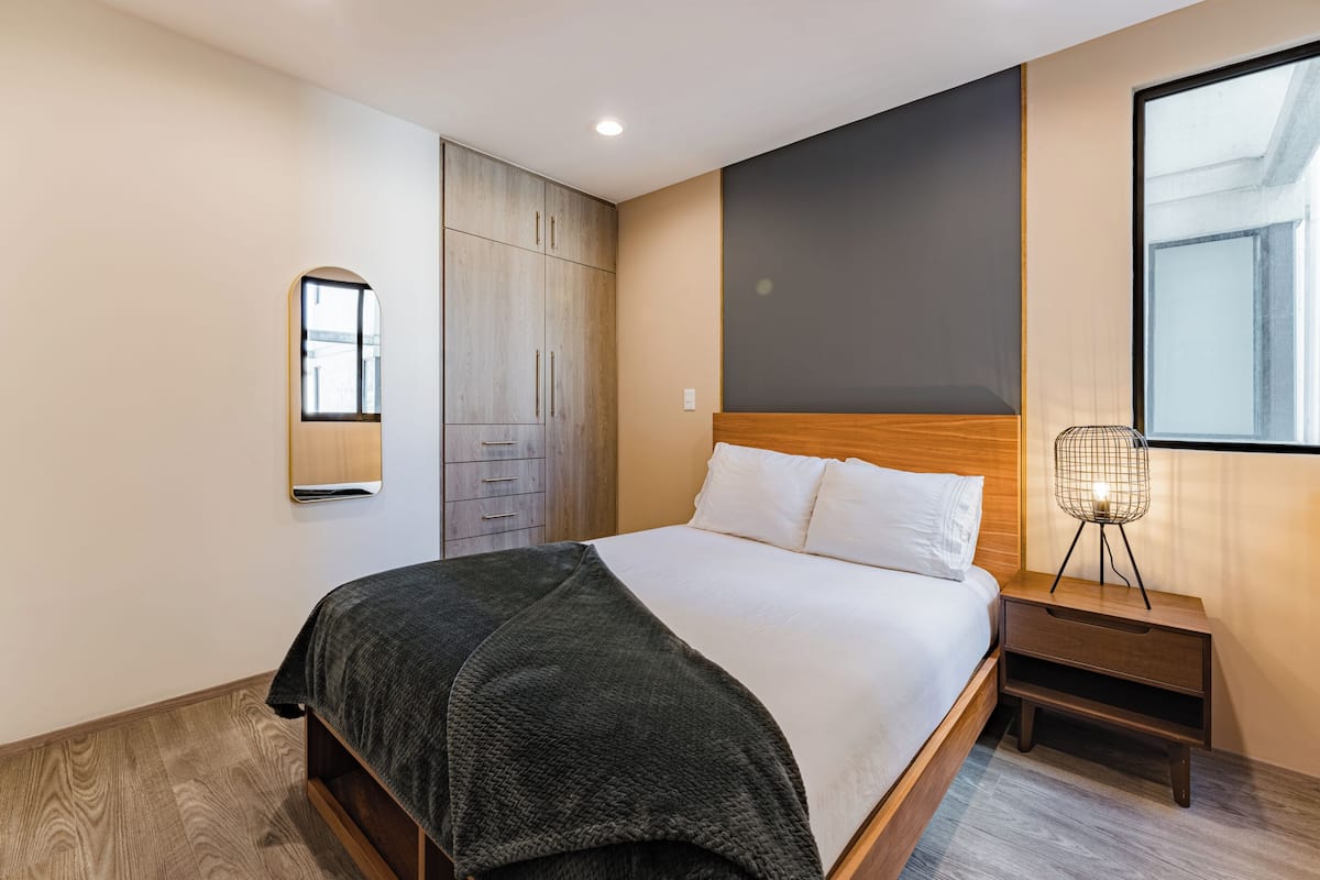 Een moderne slaapkamer met een houten bed, een nachtkastje met een lamp, een garderobe en een groot raam.
