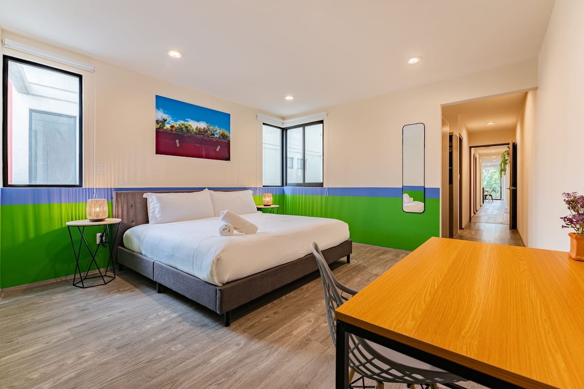 Een moderne hotelkamer met een bed, een houten tafel en een stoel. Er hangt een schilderij aan de muur en er is een raam met jaloezieën.