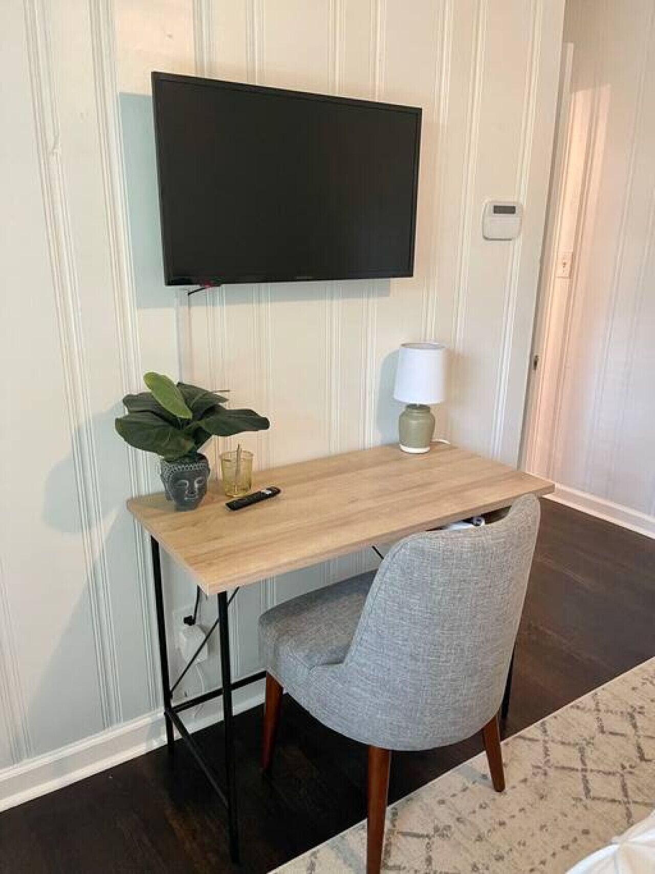 A small, neatly arranged workspace with a wooden desk, a grey chair, a potted plant, a remote control, and a wall-mounted TV.