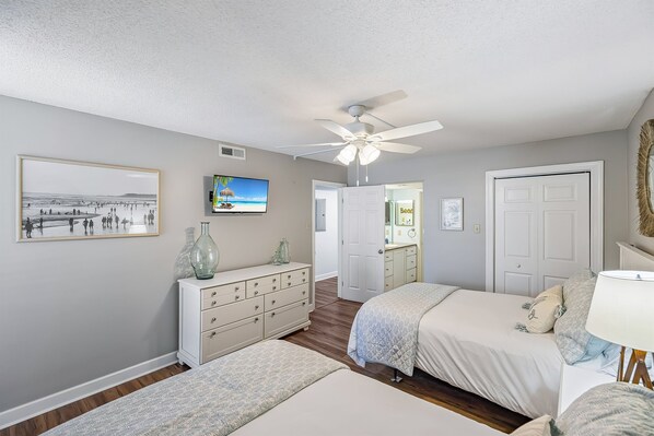 A bedroom with two beds, a ceiling fan, a TV, a dresser, and a framed picture on the wall. - PCB Sunkissed is beachfront to enjoy sounds of the surf and sunsets (Panama City Beach)