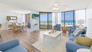 Ein geräumiges Wohnzimmer mit großem Fenster mit Blick auf den Strand, einem Essbereich mit Korbmöbeln und einem Deckenventilator.