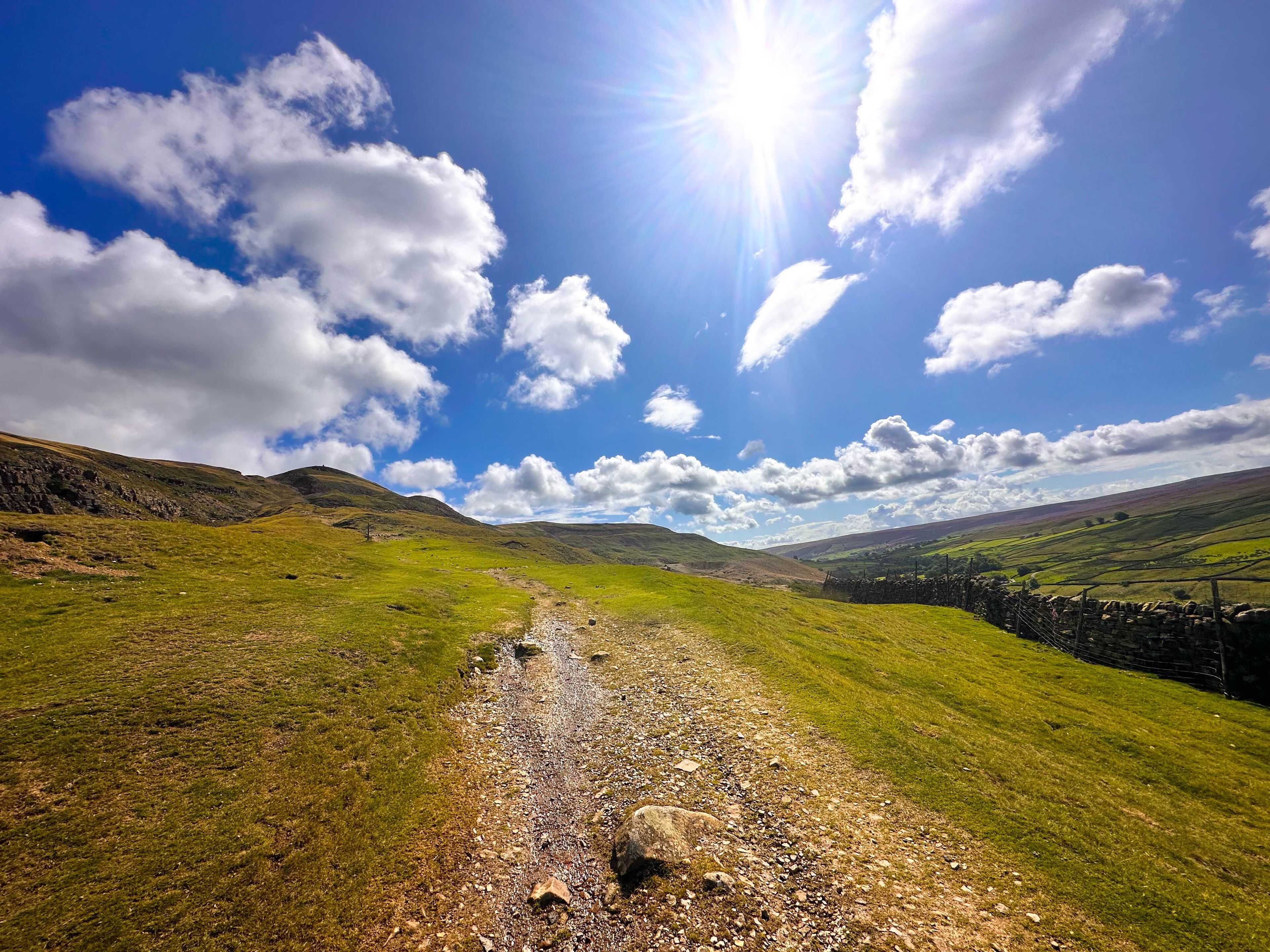 A clear sky with the sun shining, a grassy path, and rolling hills in the distance.