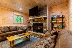 A cozy cabin interior with a stone fireplace, wooden walls, a leather sofa, and a television mounted above the fireplace.