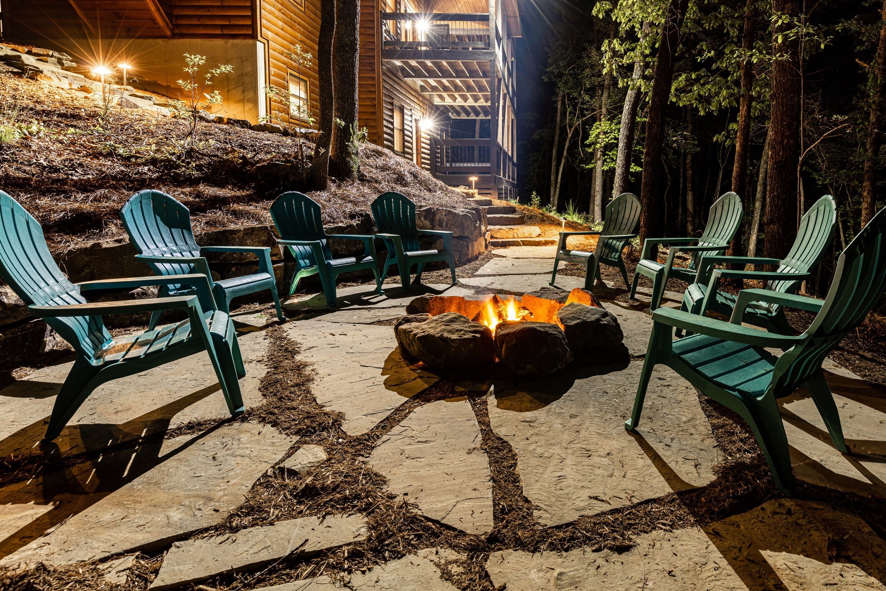Een vuurkorf van steen, omringd door groene Adirondack-stoelen, in een openlucht omgeving met houten gebouwen op de achtergrond.